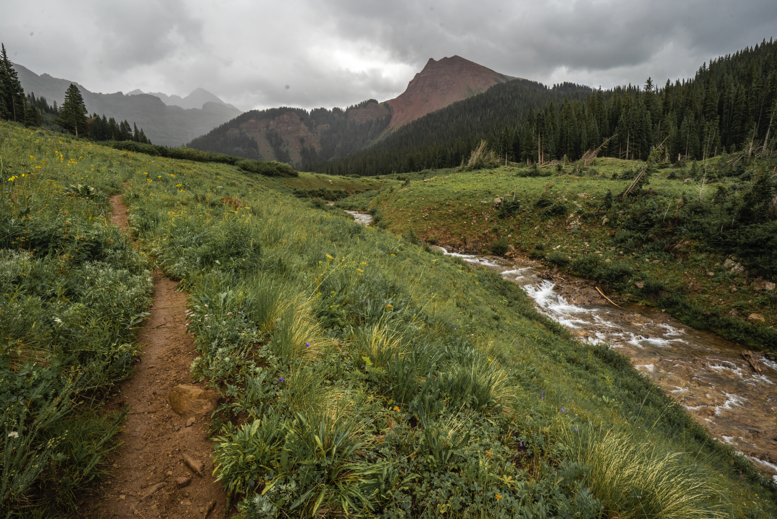 Trail moves next to a stream with large mountains looming in a foggy background