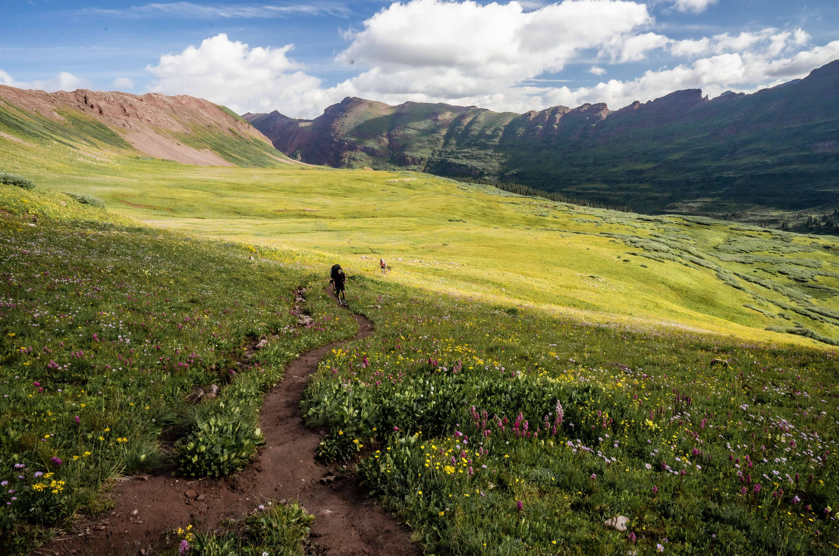 A hiker hikes towards the photographer with a sunny meadow and mountains in the background